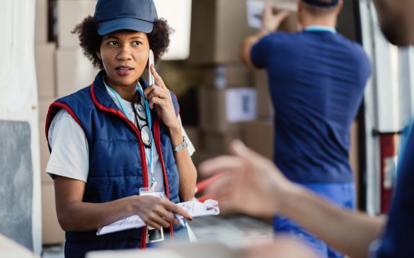 Black female deliverer communicating with coworker while talking on mobile phone and organizing package delivery.